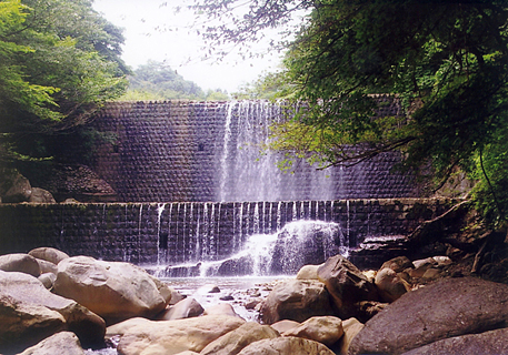 ごつごつとした岩が沢山ある川の奥に見えている荒川第六堰堤の写真