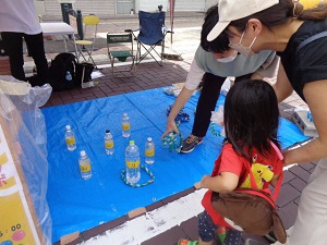 ビニールシートに並べられたペットボトルに向かって輪投げをしようとしている女の子の後ろ姿の写真