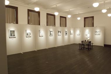 濃ゆい茶色のフローリングで白壁をした室内で、壁際のパネルに等間隔に作品が展示されている展示室内の写真
