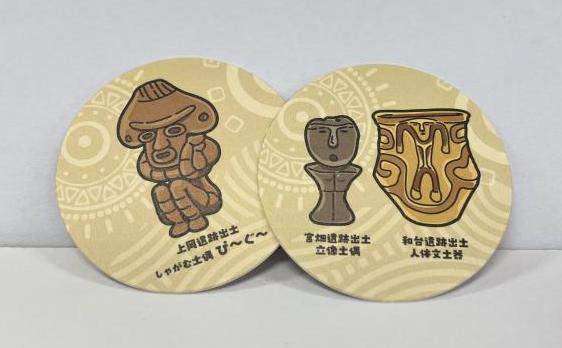しゃがむ土偶が描かれたコースターと立像土偶と土器が描かれたコースターの写真