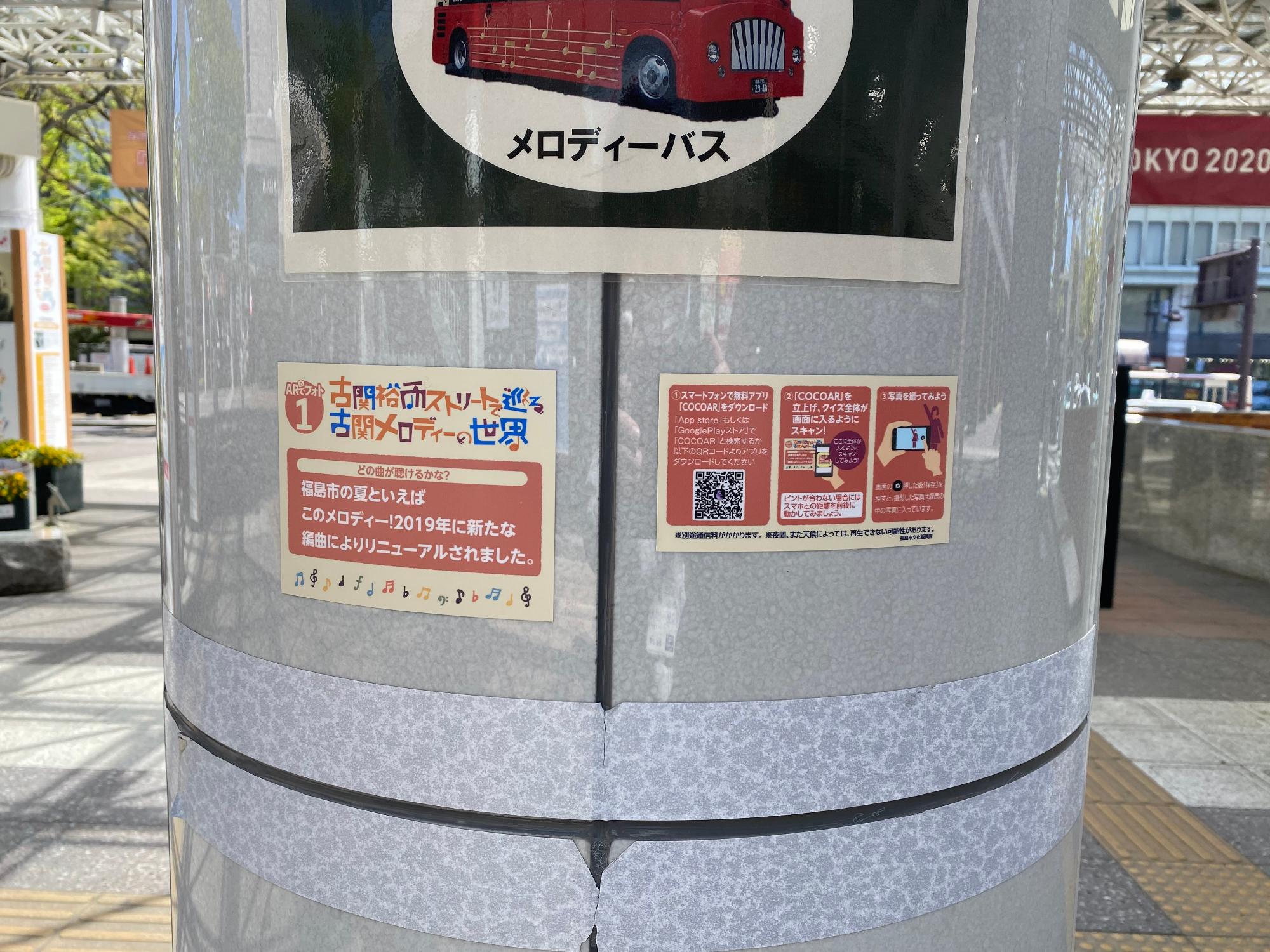 屋外にある円柱状の柱に「古関裕而ストリートで巡る古関メロディーの世界」と書かれたポップのような物が貼られtている写真