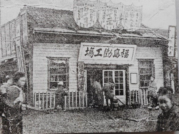 人が多く行き交う道沿いに「福島福島勧工場」と書かれた看板が出入口ドアの上に設置されている勧工場の白黒写真