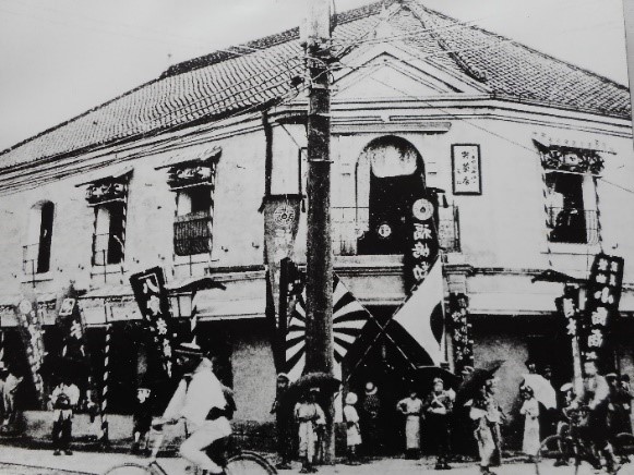 日本国旗や店ののぼり旗などが周りにたくさん立てられた2階建ての勧工場の建物前に大勢の人が集まっている様子の白黒写真