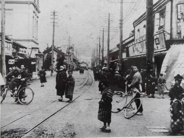 店が立ち並び中央に線路が通った大通りがたくさんの人で賑わい、自転車に乗っている人たちがいる様子の白黒写真
