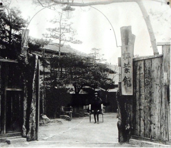 「松葉館」と書かれた木の看板が設置された門の奥に、植栽と建物がみえている明治時代の松葉館を撮影した白黒写真