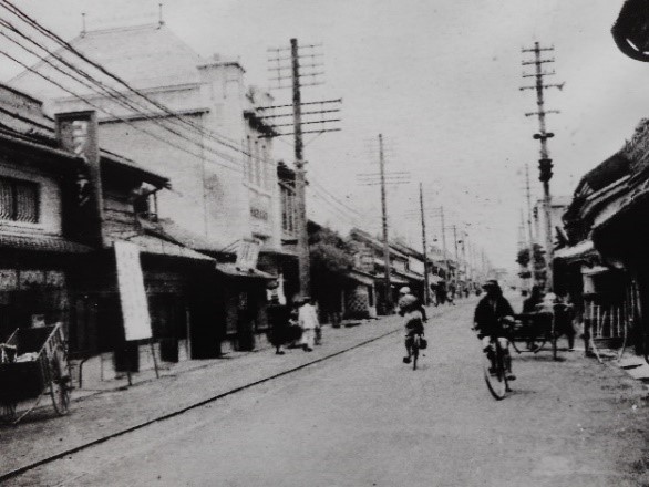 商店が軒を連ねる通りには、自転車を走らせる人や行き交う歩行者が見られ、道端には荷車が置かれ、通りの左手には重厚な洋風建築がある大町通りを写した白黒写真