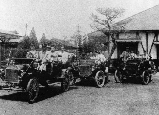 制服を着ている運転手がハンドルを持ち、着物姿の女性や男性が数名ずつ乗っているオープンカーの乗合自動車が3台写っている白黒写真