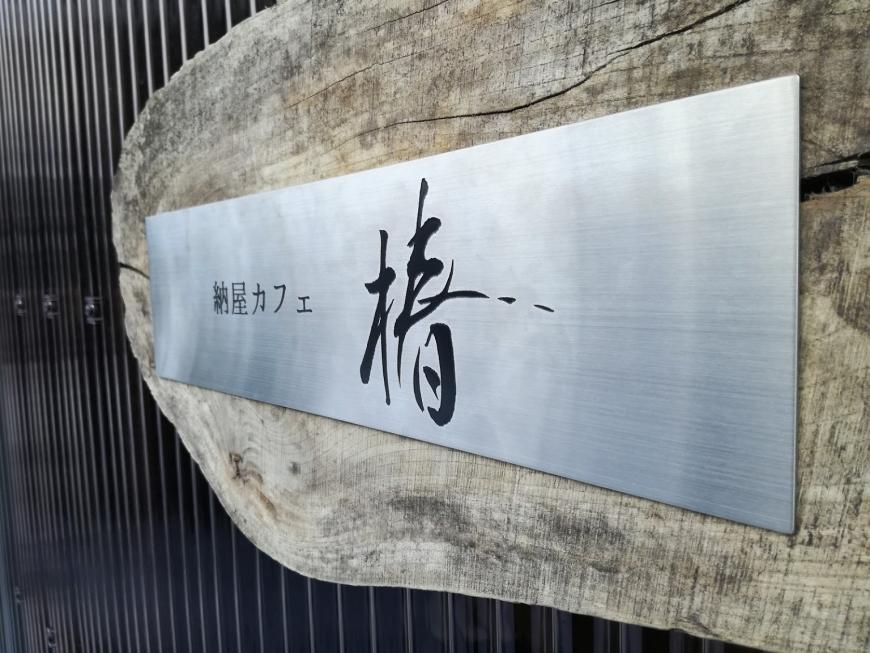 木の板に貼られたシルバーのプレートに黒字で「納屋カフェ椿」と書かれている看板の写真