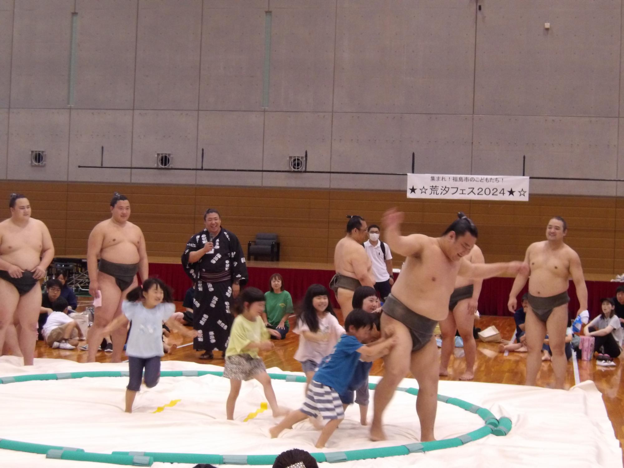 5人の子供たちが土俵に上がり、そのうちの2人の子供たちが大きな力士を土俵際に追いやっている写真