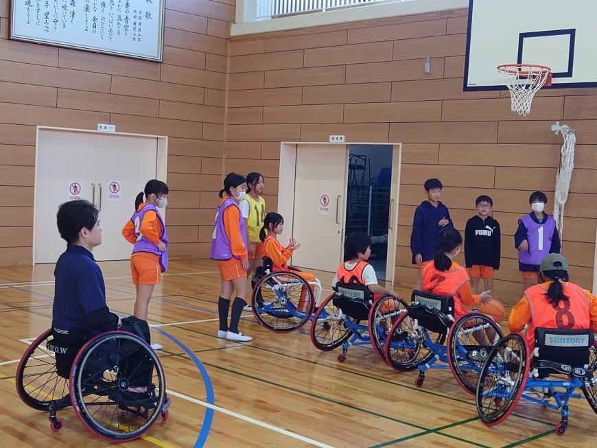 バスケットリングの前に車いすに乗り横一列に整列している4人の生徒達を後ろから見守っている豊島英氏の写真