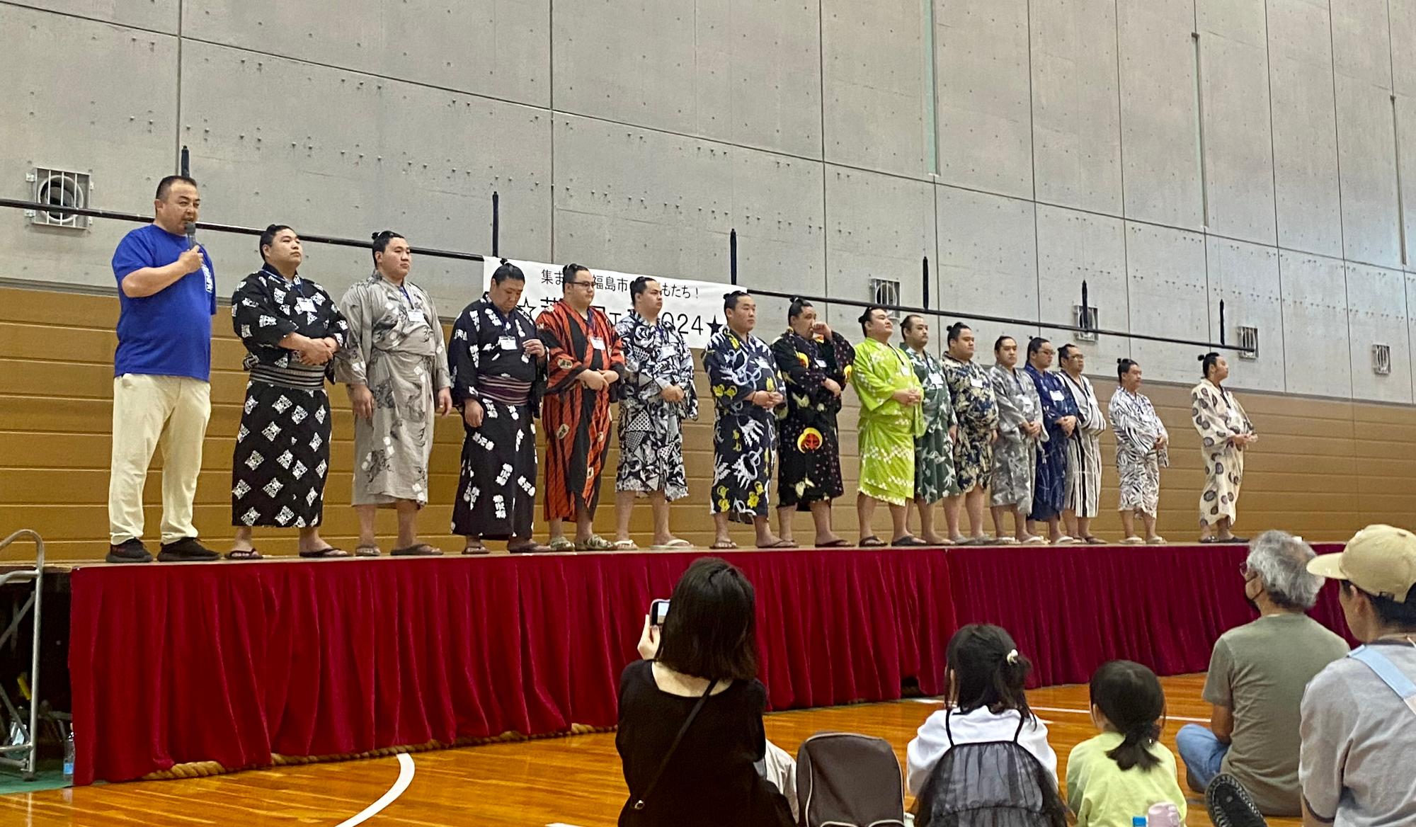 15名の力士と荒汐親方がステージ上に横一列に並び、マイクを持った荒汐親方が集まった参加者達に向かって話をしている様子の写真