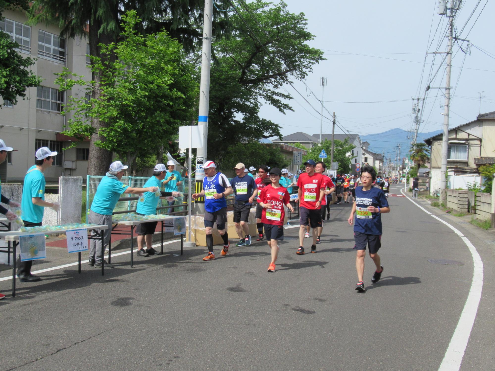 沿道に長机を設置し、エメラルドグリーンのシャツを着た関係者の方々がランナーに何か手渡そうと手を伸ばしている写真