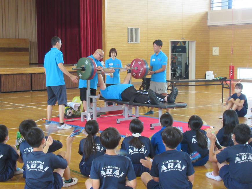 渡邉和幸選手がベンチ台に寝てバーベルを持ち上げている様子を生徒達が見ている写真