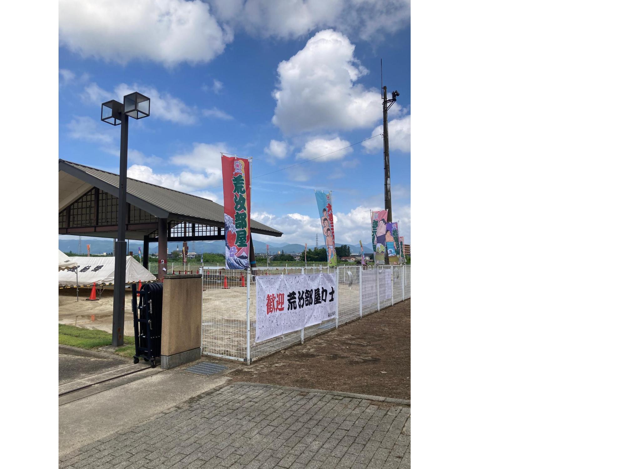 広場のような場所の入口門の横に「荒汐部屋」と書かれたのぼり旗があがっていたり、門の横のフェンスには「歓迎荒汐部屋力士」と書かれた横断幕が掲げられている写真