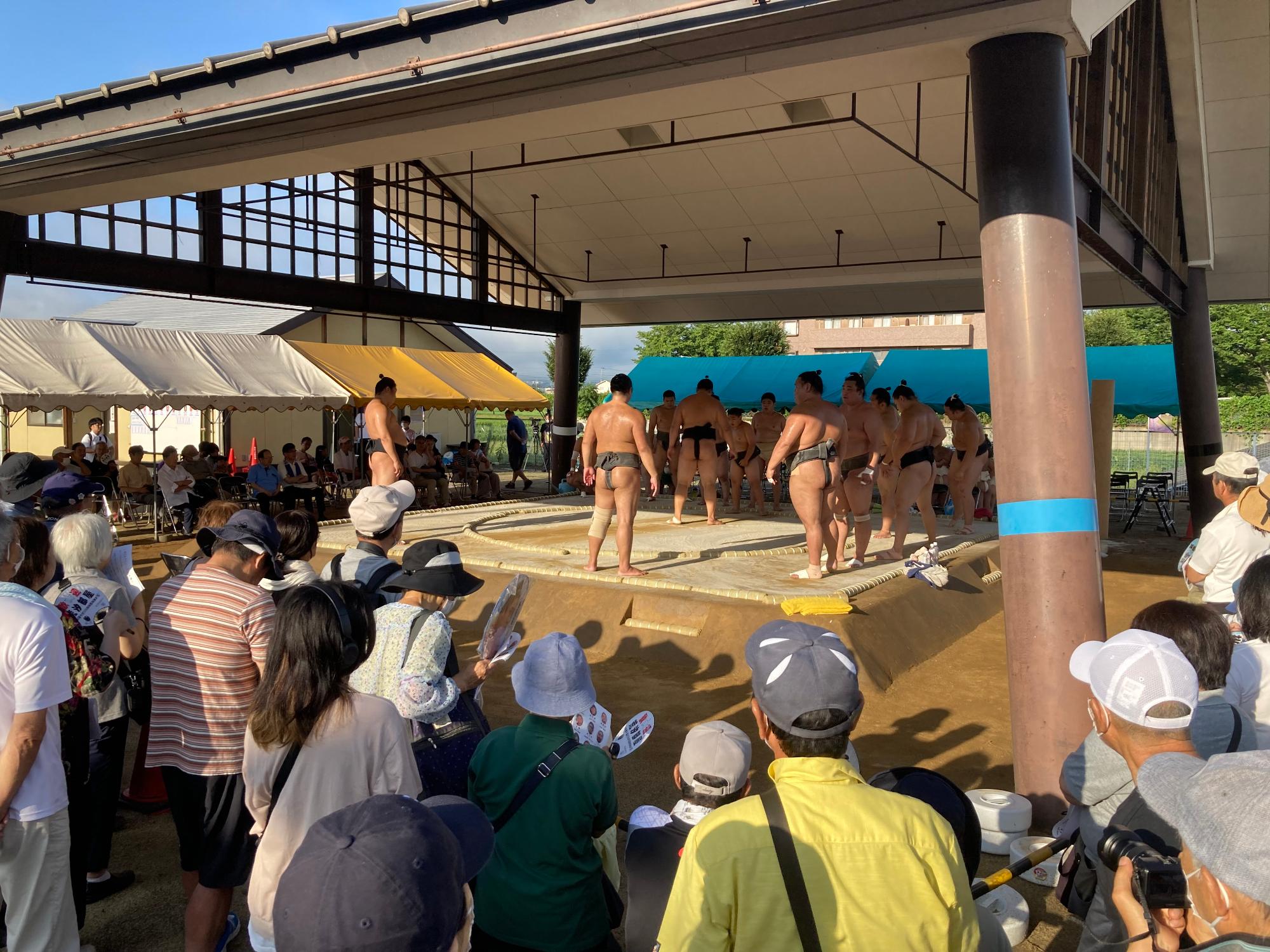 屋外の屋根付きの土俵場にふんどし姿の力士たちが間隔をあけて立ち、その様子を多くの方が見ている写真