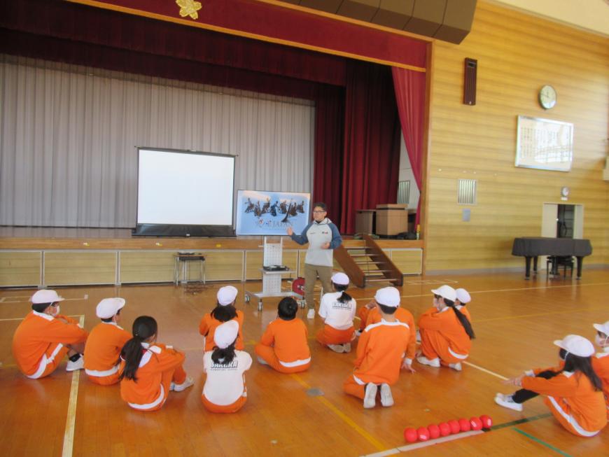 体育館ステージ側に設置されたスクリーンとモニターの横に村上光輝氏が立ち、床に座っている子供たちに話をしている様子の写真