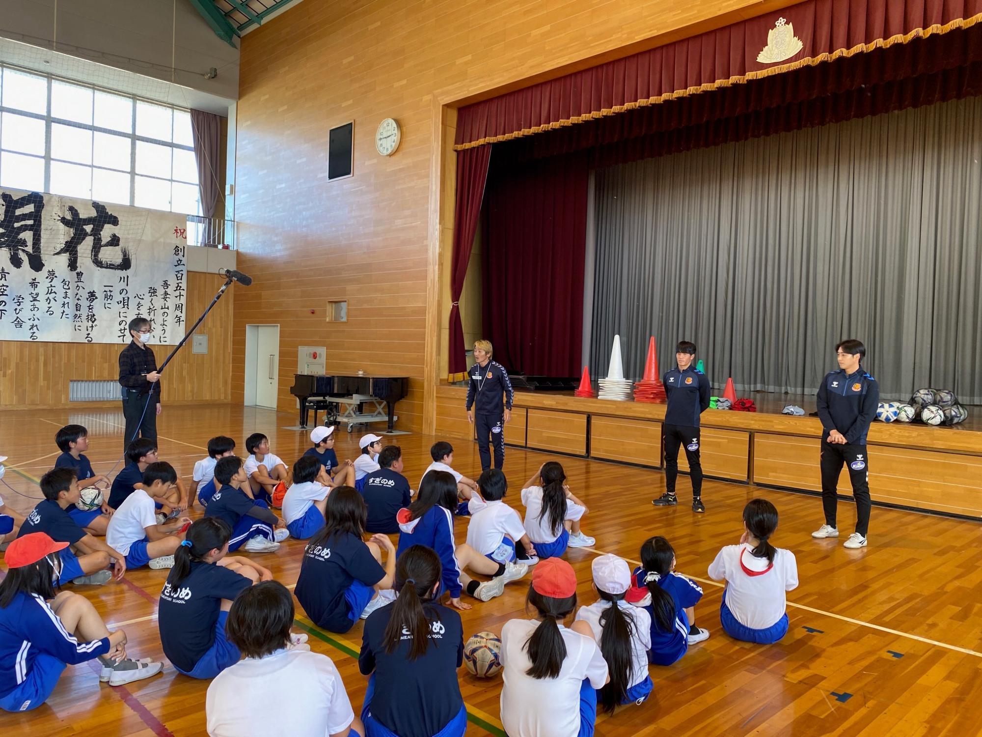 体育服を着た生徒達が体育館の床に座り、3人の選手たちが子供たちに出前授業を行っている様子の写真