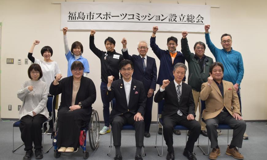 「福島市スポーツコミッション設立総会」と書かれた横断幕が掲げられている前で、市長を中心に関係者達が集まり、右手で握りこぶしを作り腕を上に上げるポーズで記念撮影をしている写真