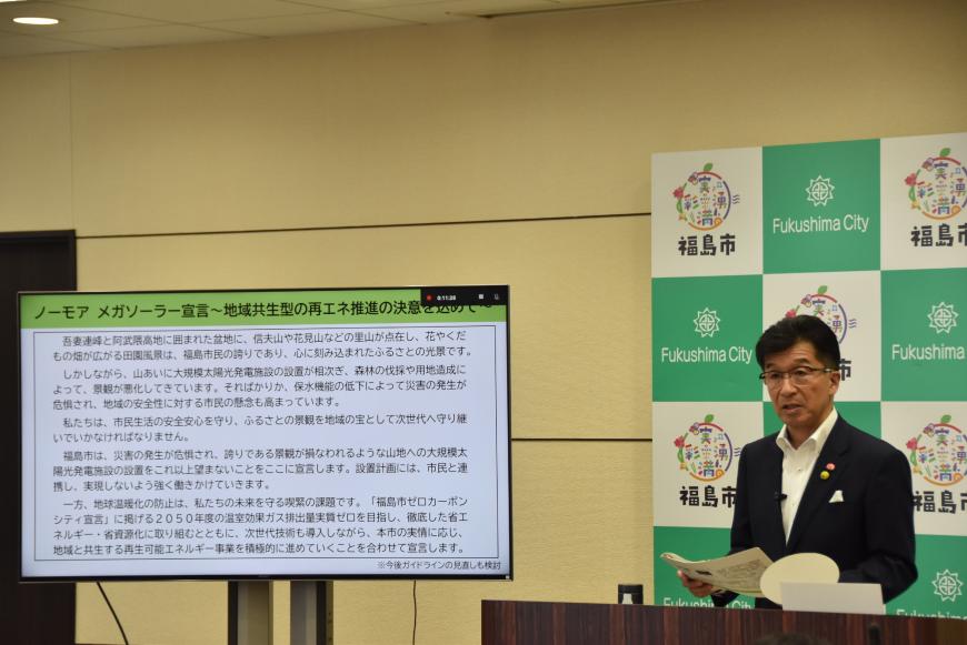 ノーモア メガソーラー宣言についての資料が映されたモニターの横で記者会見を行っている市長の写真