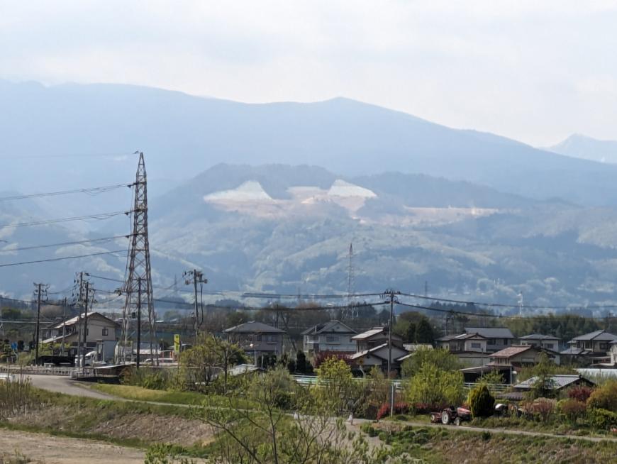 左側に鉄塔が立ち民家が立ち並んだ奥に一部山肌が見えている先達山の写真