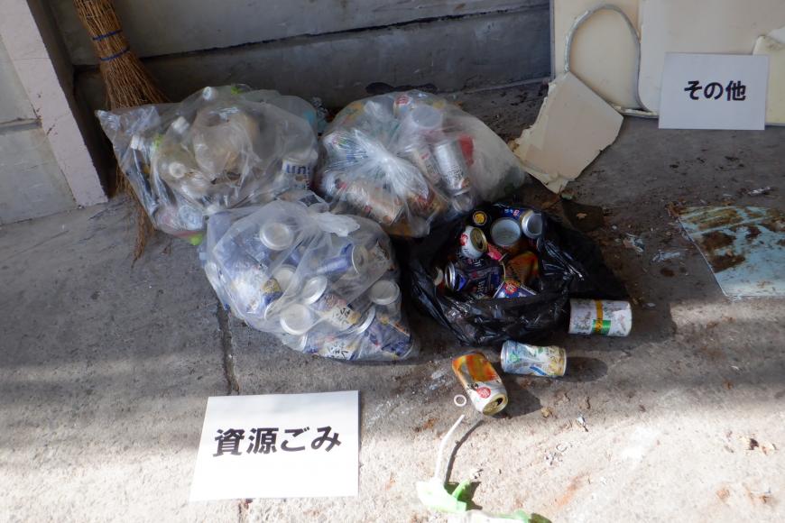 「資源ごみ」と書かれた用紙が置かれペットボトルや不燃缶が透明袋や黒色のゴミ袋に沢山入っている写真