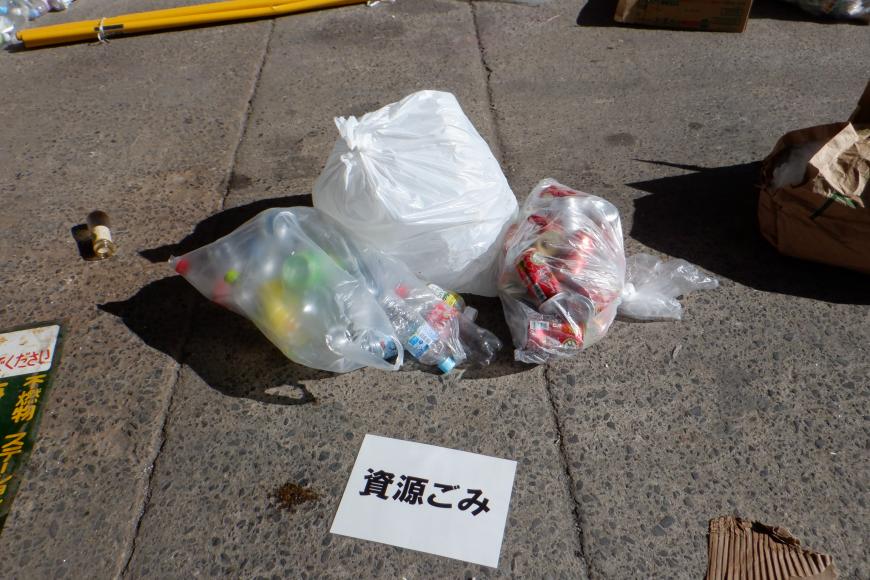 「資源ごみ」と書かれた用紙が置かれ、袋に入ったった大量のペットボトルや缶が3袋置かれている写真