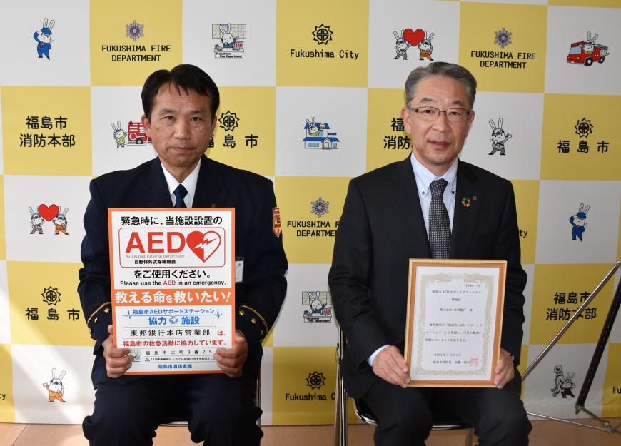 事業所設置掲示板を持っている安藤消防長と盾を持っている遠藤常務執行役員本店営業部長の写真