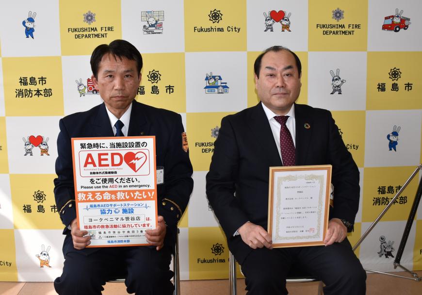 消防の制服を着た消防長がAEDのチラシ、矢吹執行役員が賞状を持ち、並んで座って記念撮影をしている写真