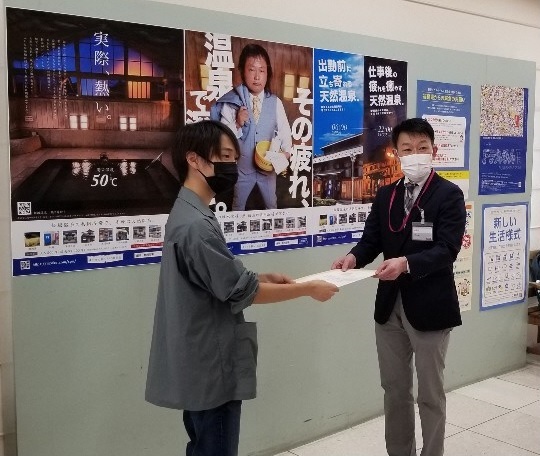 飯坂温泉PRポスターの前で関係者の男性とイオンの担当者が包括連携協定書をもっている写真