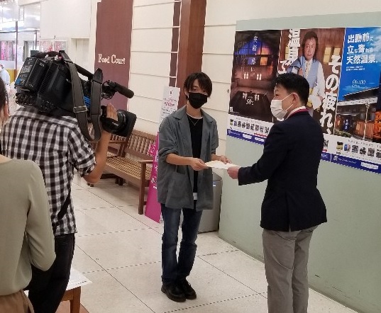 飯坂温泉PRポスターの前で関係者の男性とイオンの担当者が包括連携協定書をもっている様子をカメラマンが撮影している写真
