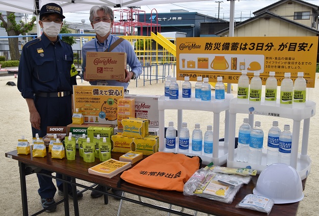紺色の制服姿の市長と補助食品が入ったダンボールを持った野崎課長補佐が並んだ屋外に設置されたテントの下に、アクエリアスなどのペットボトルが3種類、パウチやヘルメットなど防災備蓄の品物が机に並べられている写真
