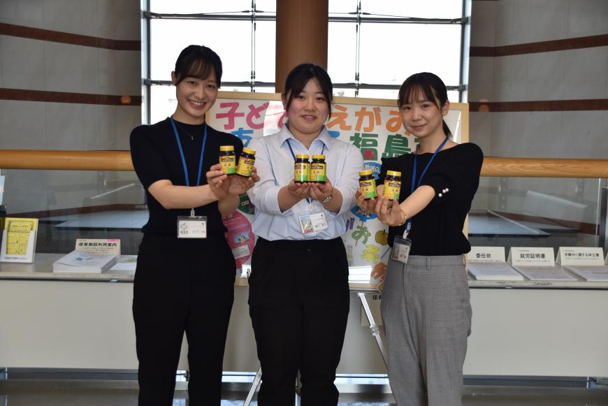 資料が置かれた台の前で、両手で葉酸サプリのボトルを持ち横一列に並んでいる女性3名の写真