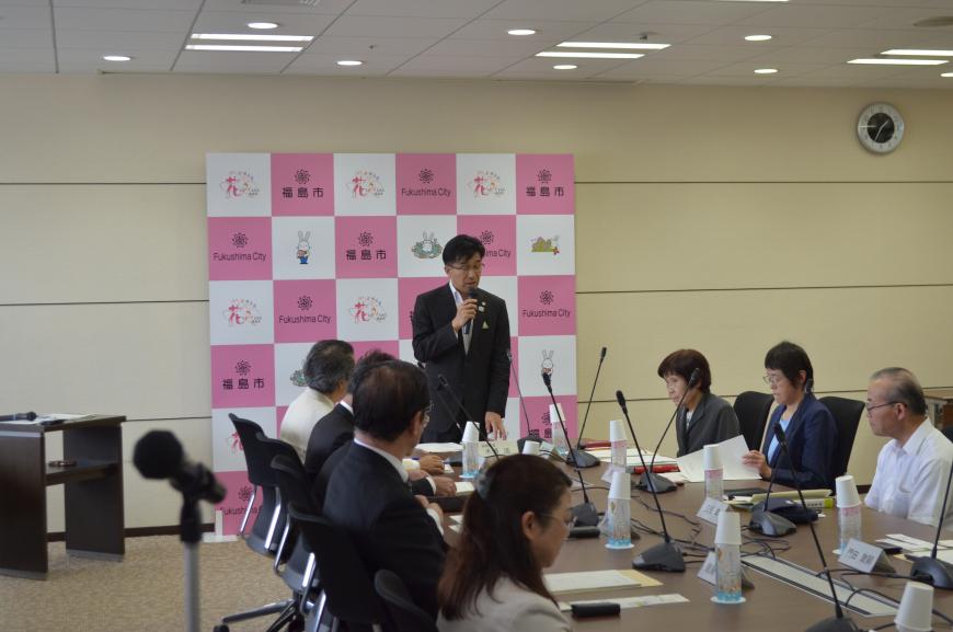 福島市のバックパネルの前に立ってマイクを持って話をしている市長と向かい合って席について話を聞いている参加者の写真