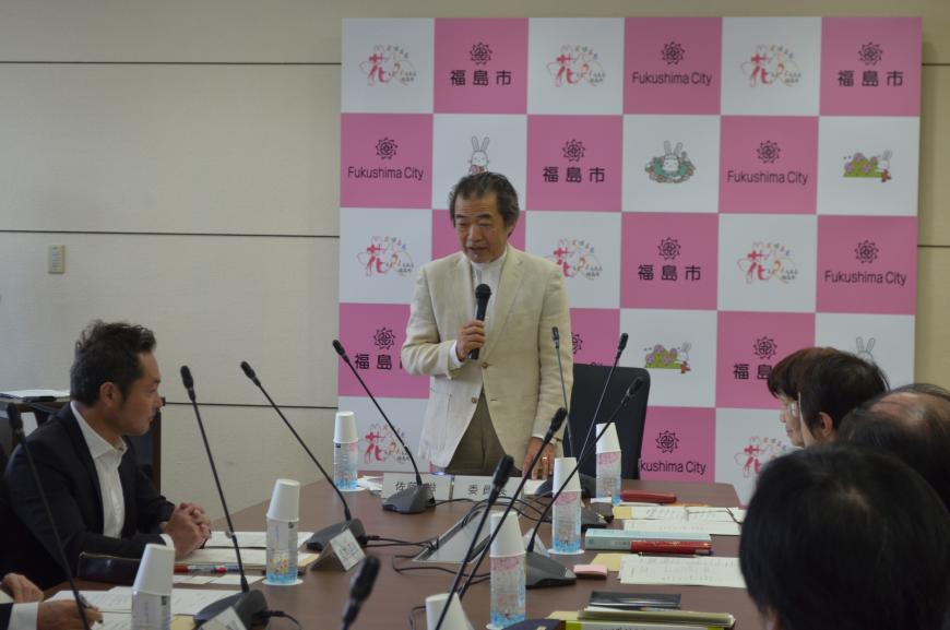 福島市のバックパネルの前に立ってマイクを持って話をしている白いジャケットの男性の写真