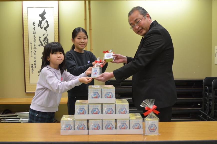 机の上に積み上げられた「ハト缶」と女の子2名が男性に「ハト缶」を受け取っている様子を写した写真