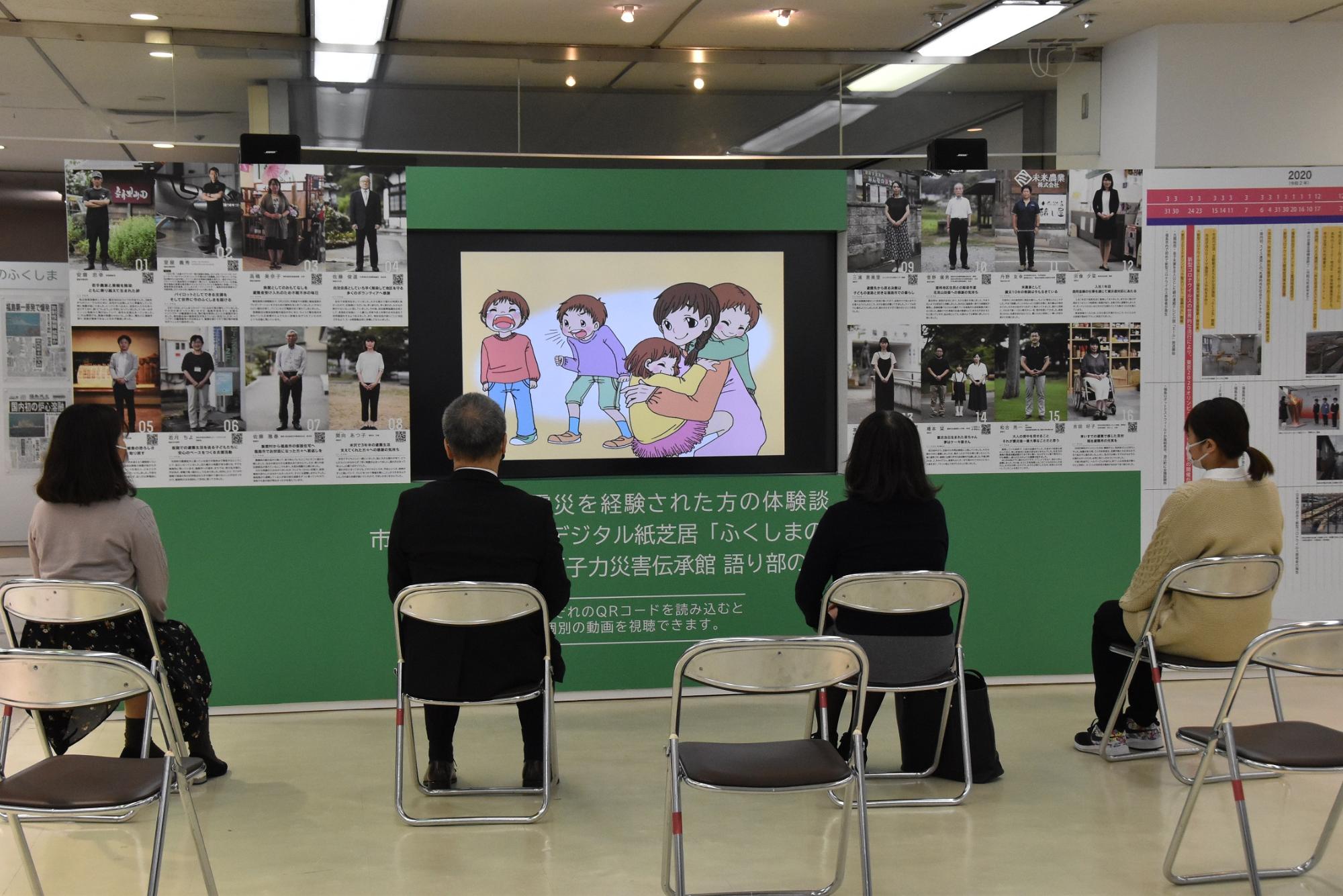 災害伝承館のモニターを4名の人がパイプ椅子に座って見学している後姿を写した写真
