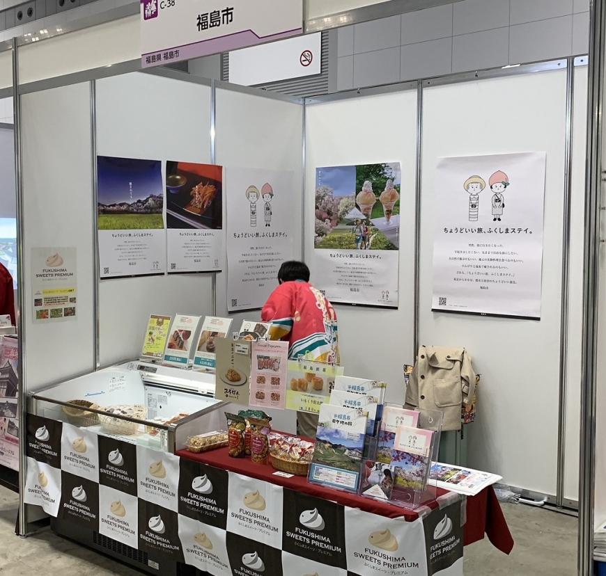 資料が並び、壁にはポスターも展示されている福島市のブースで法被を着た女性が奥で準備している様子を写した写真