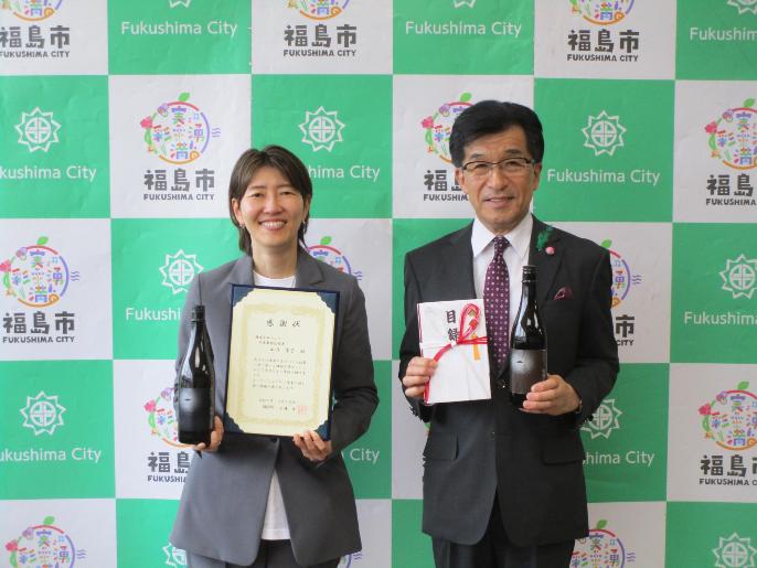 企業版ふるさと納税の感謝状の贈呈時の写真です。左に株式会社サンワの山川景子社長が感謝状と飯野の日本酒「宙飛ぶ宴絆」を持ち、右に福島市長が目録と飯野の日本酒「宙飛ぶ宴絆」を持っています。