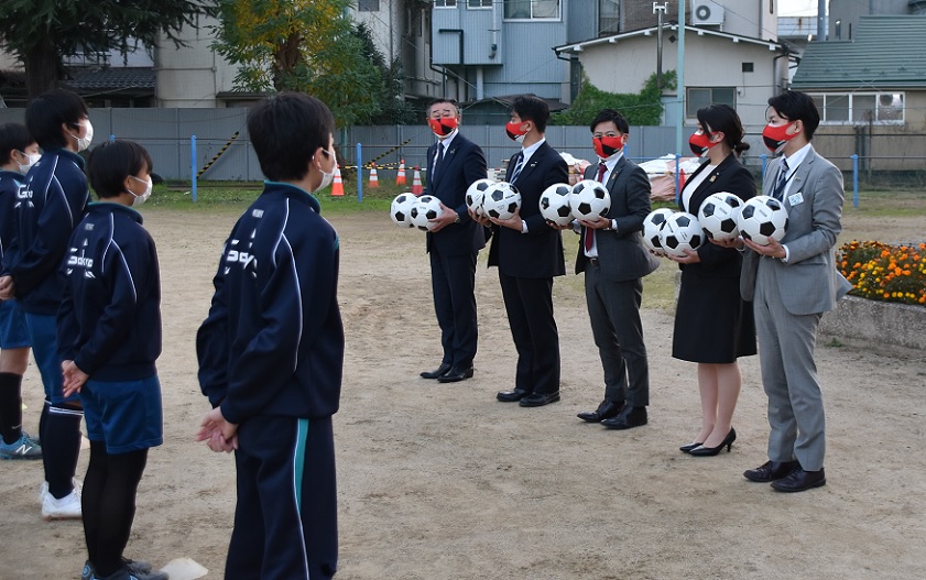 運動場に1列に並んで両手にサッカーボールを持っているスーツ姿の男女5名と対面に並んでいるジャージ姿の子供たちの写真