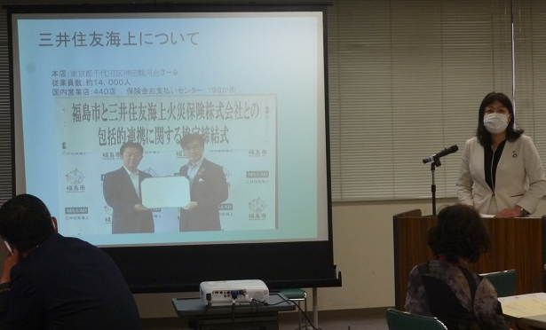 女性が演台でマイクを使い、資料が映し出されたスクリーンの横で講演をしている写真