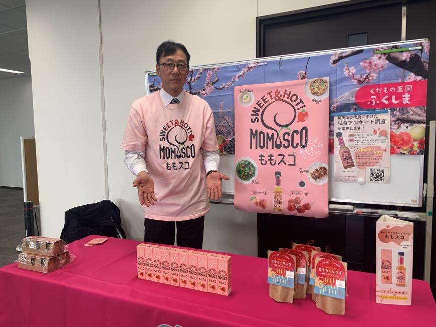 「ももスコ」と書かれたピンク色のオリジナルTシャツを着た関係者の男性が「ももスコ」などの商品が並べられたテーブルに向かって紹介するように両手のひらを広げている写真