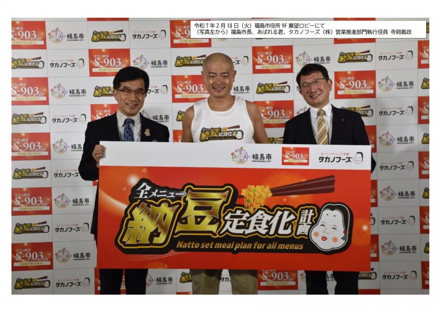 福島市とタカノフーズ株式会社のバックボードの前で「全メニュー納豆定食化計画」と書かれたボードを一緒に持ったあばれる君を中心に関係者2名の男性が記念撮影を行っている写真