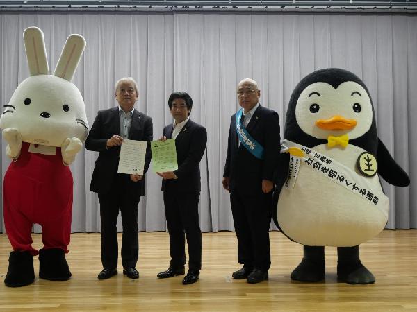 メッセージが書かれた紙を持った関係者3名と「ホゴちゃん」と「ももりん」が1列に並び記念撮影を行っている写真