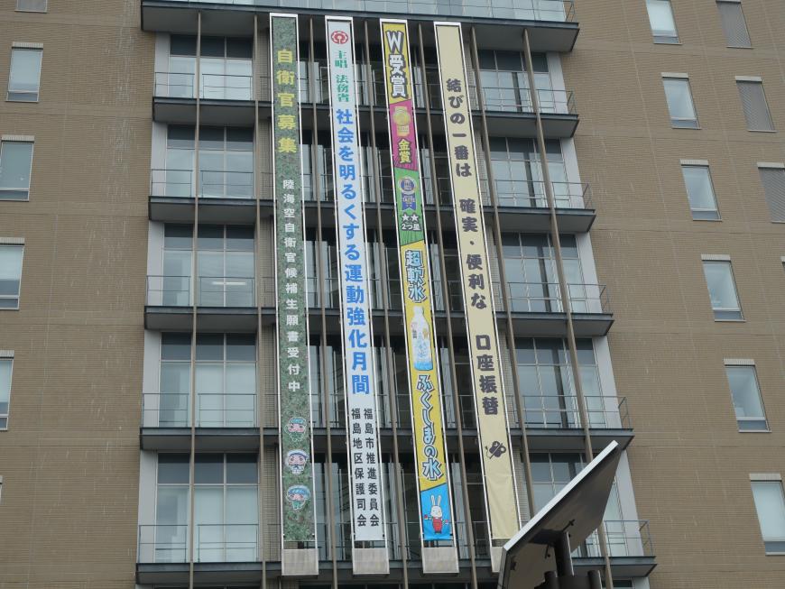 福島市役所の壁面に大きな4つ懸垂幕が設置されている様子を見上げるように撮影した写真