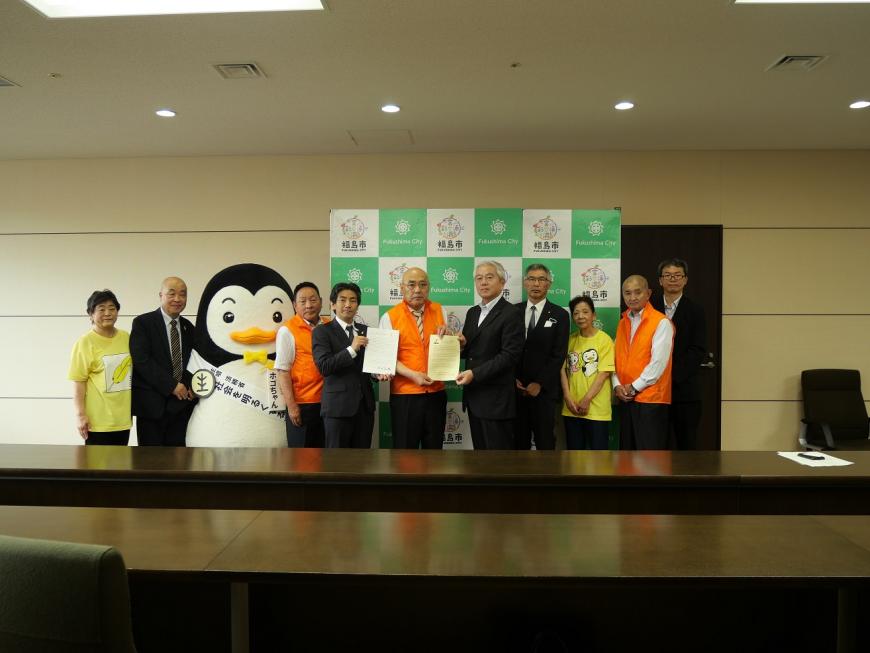 福島市のバックボードを背景にメッセージが書かれた紙を持った関係者3名と7名の関係者、ゆるキャラ「ホゴちゃん」が1列に並び記念撮影を行っている写真