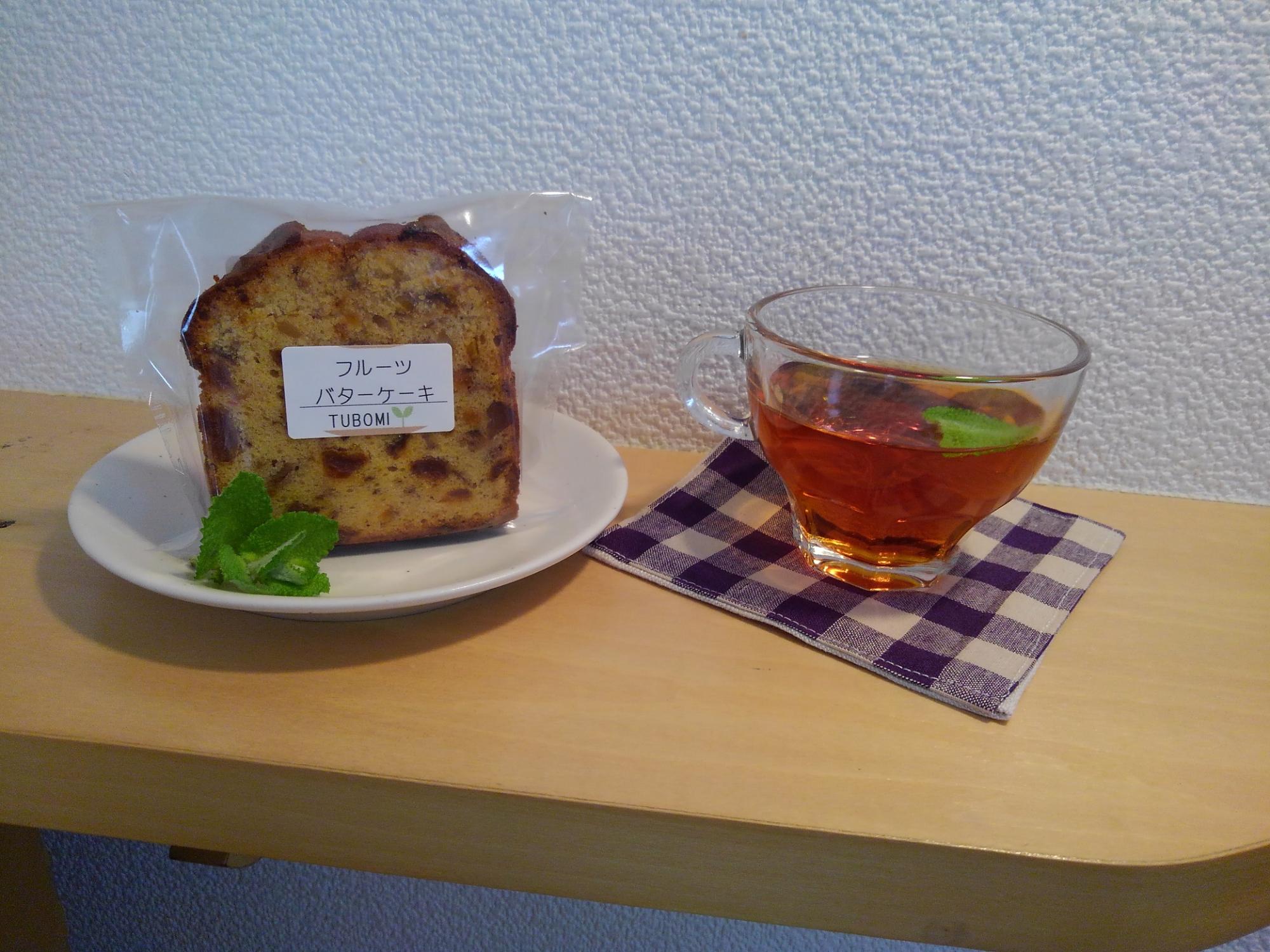 白と紫色の布のコースターの上に透明のグラスに注がれたお茶と、白皿の上に透明の袋に入ったフルーツバターケーキが置かれた写真