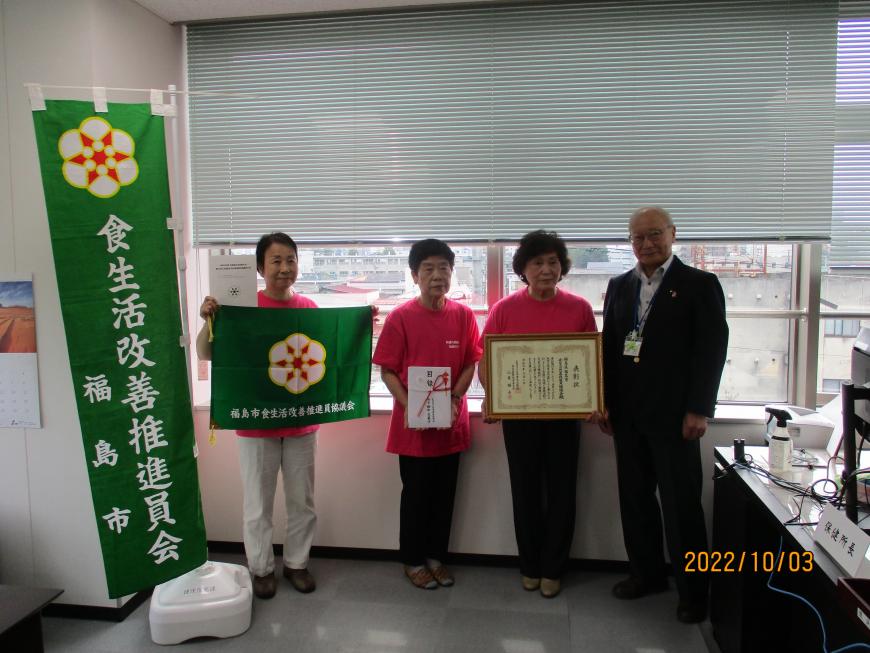 緑色の「食生活改善推進員会」ののぼりの横で、のぼりと同じデザインの布を広げて持った女性、目録の封筒を持った女性、表彰状を持った女性とスーツ姿の男性が並んで写した記念写真