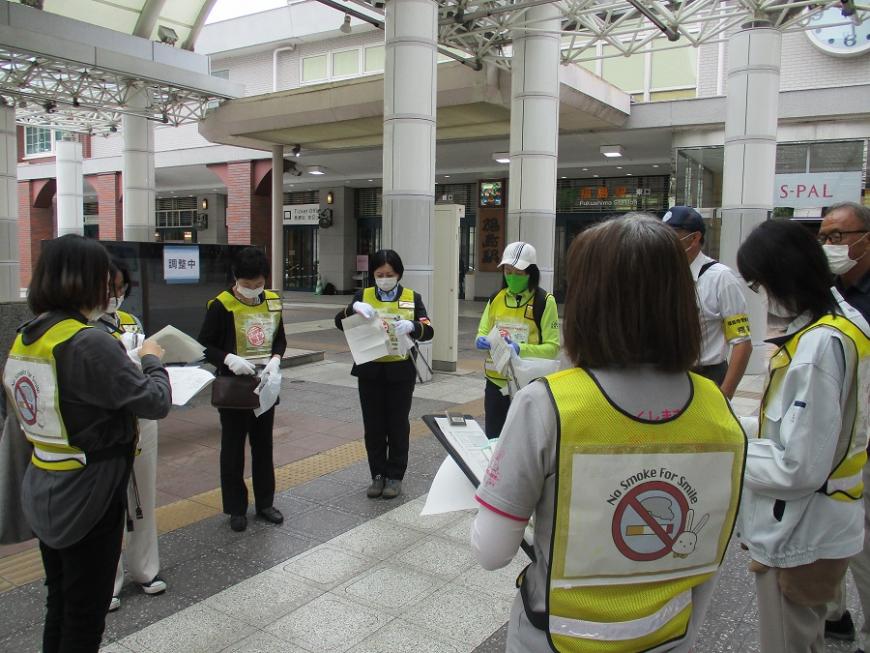 駅前のスペースに輪になって立ち、手に持った用紙を確認している黄色のビブスを身に着けた参加者たちの写真