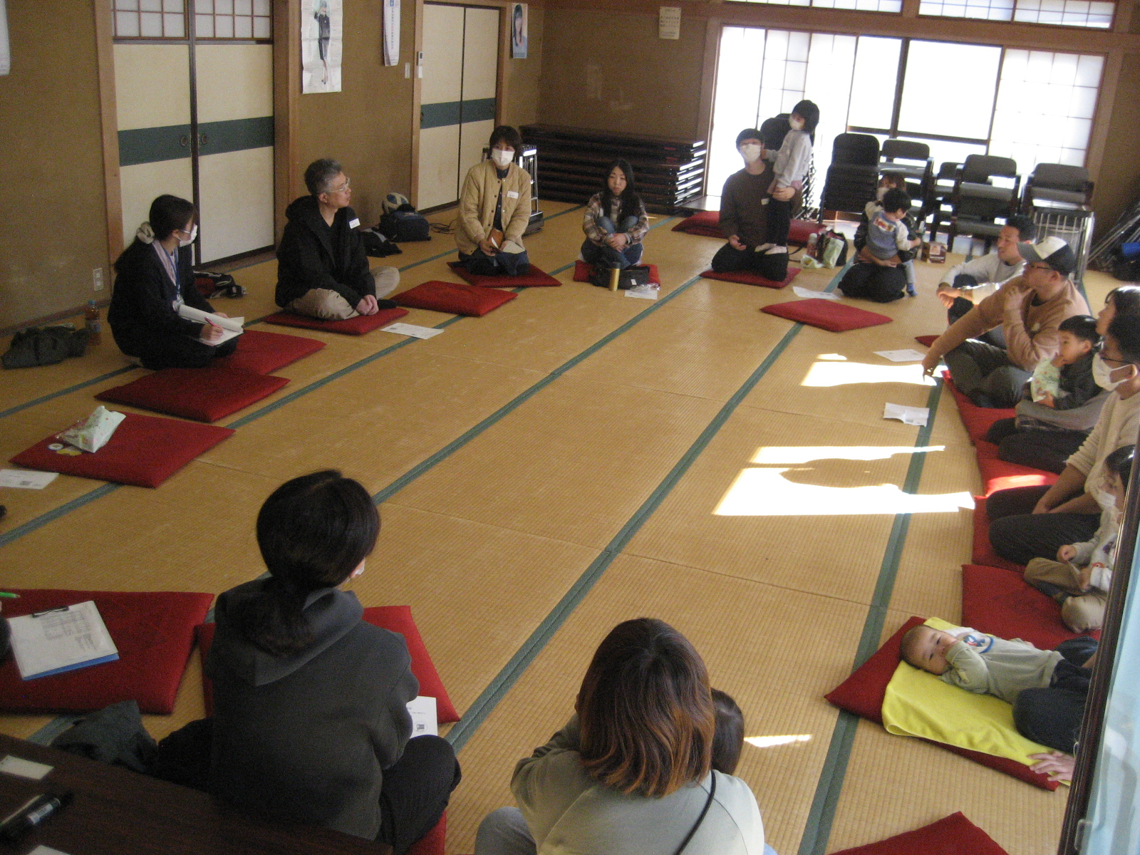 座敷に赤い座布団が丸く並べられ、小さい子供を連れた参加者たちが輪になって話をしている写真