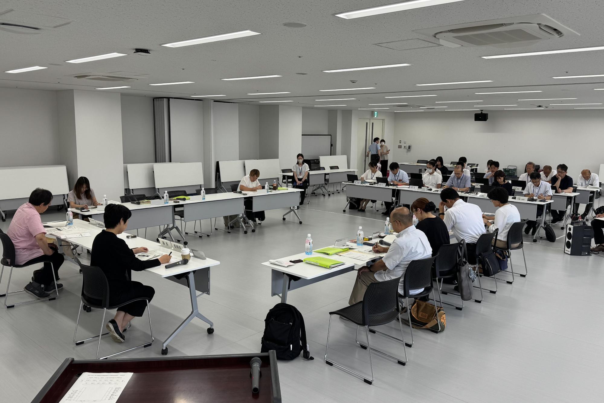 会議室にコの字型に並んだ会議机とその正面に横に数列並んだ会議机の席に出席者たちが座っている委員会の様子の写真
